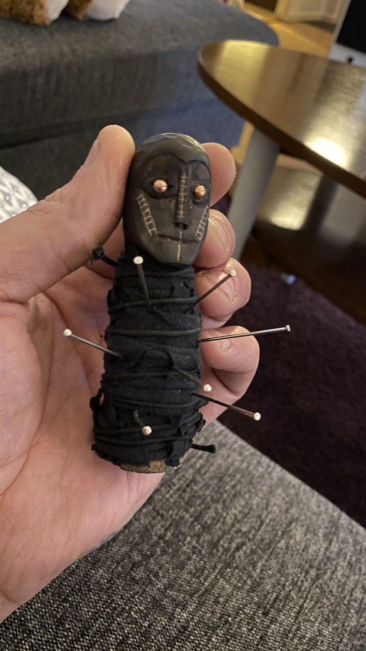 A hand holding a small carved doll wrapped in black cloth and stuck with pins